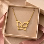 Gold-tone large butterfly outline name necklace displayed in an open beige jewellery box