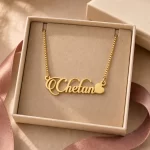 Gold-tone decorative script name necklace with a heart displayed in an open beige jewellery box