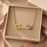 Gold-tone name necklace with a blue evil eye accent displayed in an open beige jewellery box