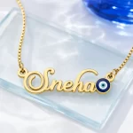Gold-tone name necklace with a blue evil eye accent on a clear glass display