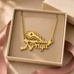 Gold-tone peacock feather and flute name pendant displayed in an open beige jewellery box