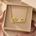 Gold-tone name necklace with crown, star and heart accents displayed in an open beige jewellery box